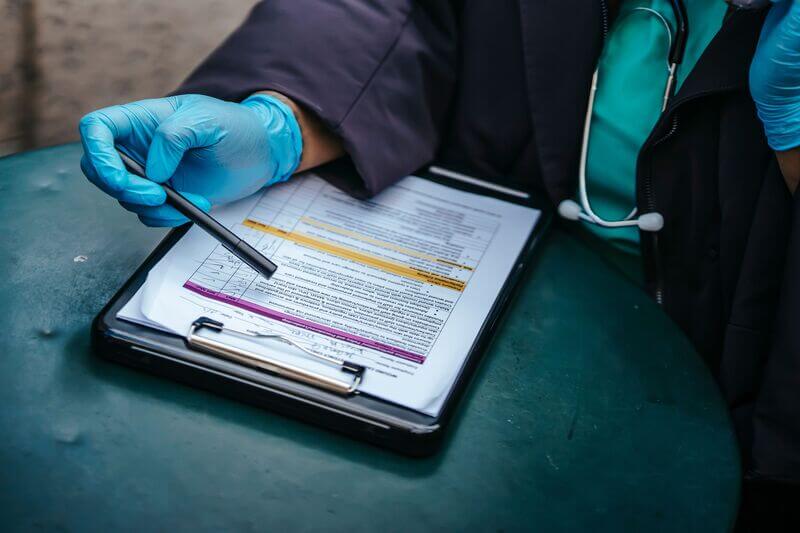 A care worker in blue gloves and a stethoscope pointing a pen at a handover form on a clipboard.