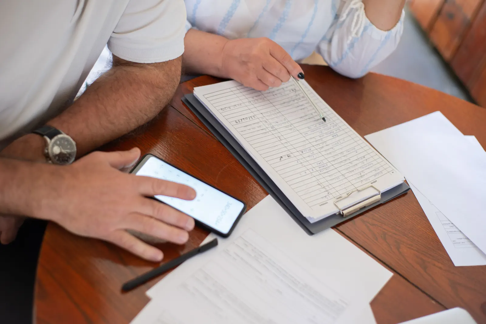 Two people at a desk going through handwritten care records on a clipboard, with a phone and paperwork beside them.