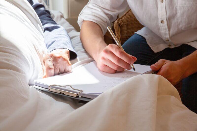 A caregiver writing notes on a clipboard beside an older resident in bed, in soft morning light.